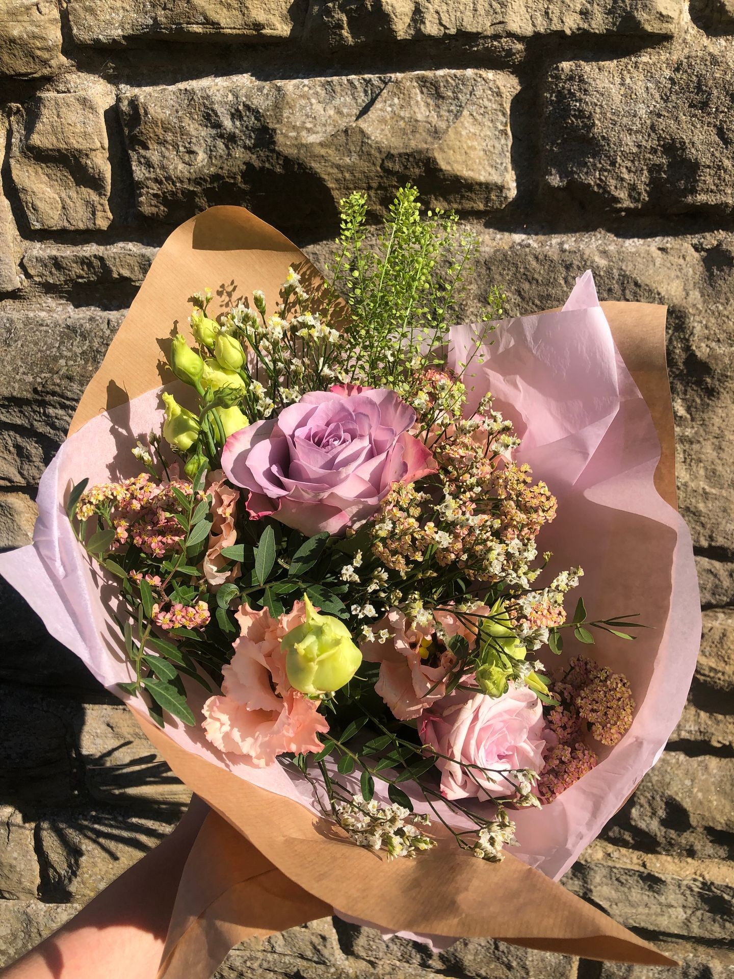 Bouquet of pink and green flowers wrapped in brown paper against a stone wall background.