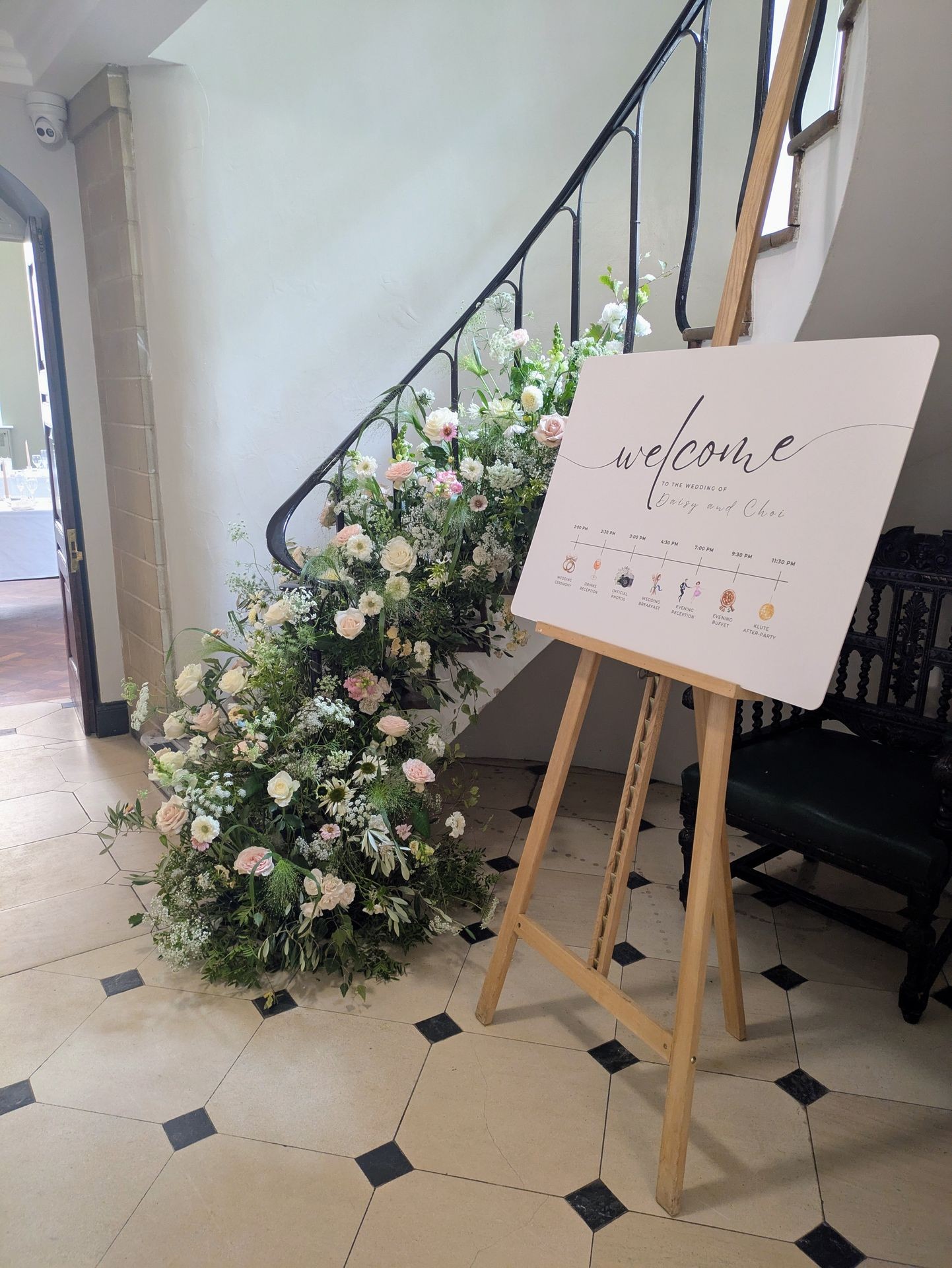 Wedding welcome sign on an easel beside a staircase adorned with lush floral arrangements.