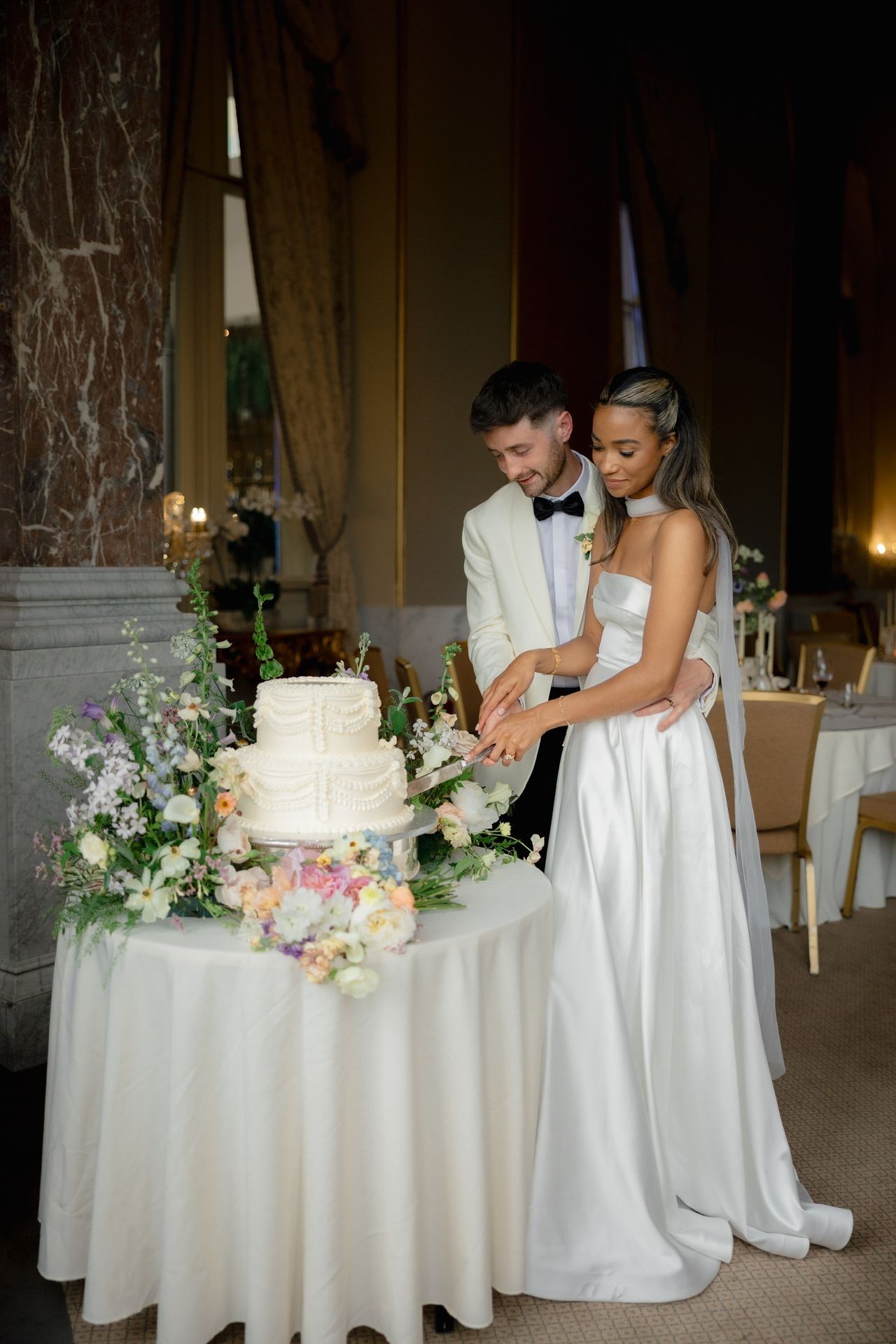 Bride and groom cutting a white tiered wedding cake adorned with flowers on a decorated table.