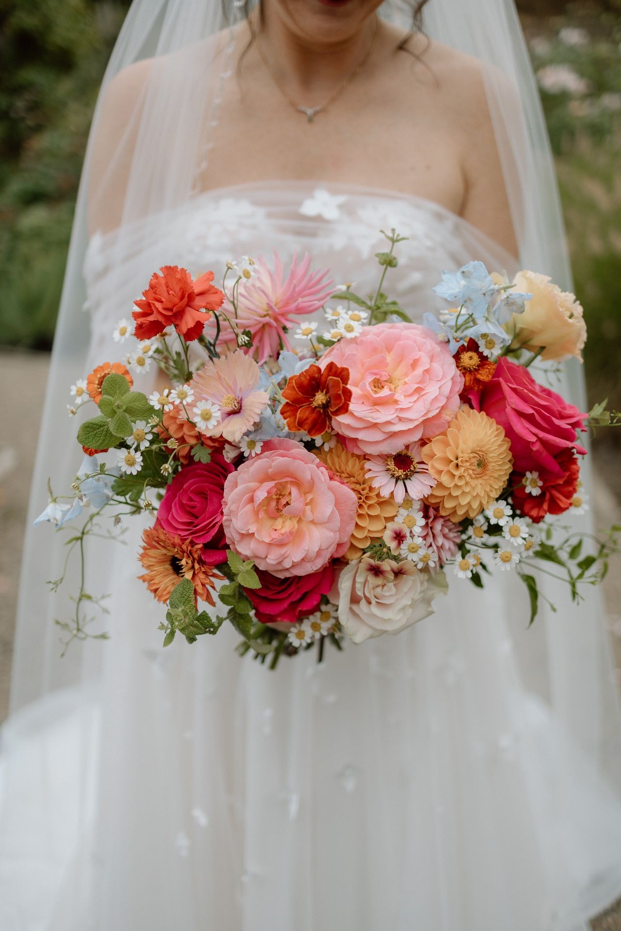 Bride in white dress holding bouquet of colorful flowers including roses, daisies, and carnations.